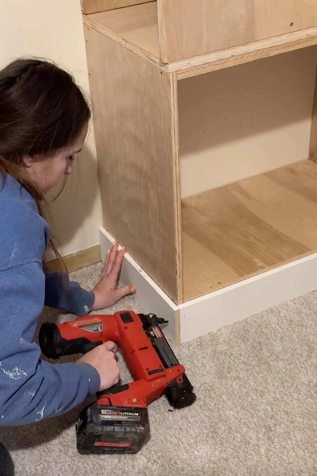 A woman in a blue sweatshirt securing white base trim to the bottom of a built-in shelving unit with a red cordless nail gun. She is aligning the trim against the carpeted floor to give the structure a finished, built-in look.